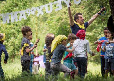 Aurélien, fondateur de Kidays, s'amuse avec des enfants joyeux dans un parc lors d'un anniversaire sur le thème Koh Lanta, les bras levés pendant que les enfants courent autour de lui.