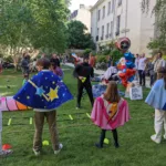 Animateur déguisé et enfant en costume fêtant un anniversaire en plein air, entourés de parents souriants qui les regardent.